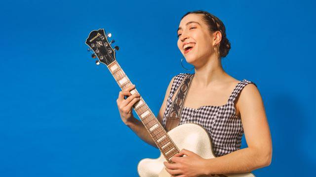 Alisa Amador smiles while singing and playing guitar. She is wearing a gingham dress and is standing if front of a bright blue background.