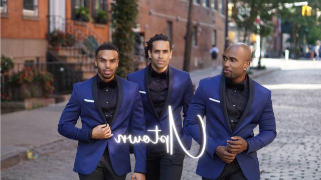 The three members of Uptown walk down a street next to a brick building. All three men are wearing blue jackets.